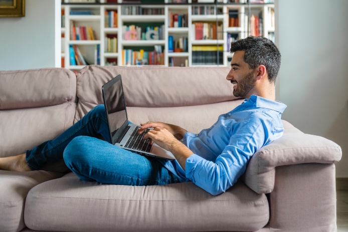 Smiling young man lying on the couch at home using laptop