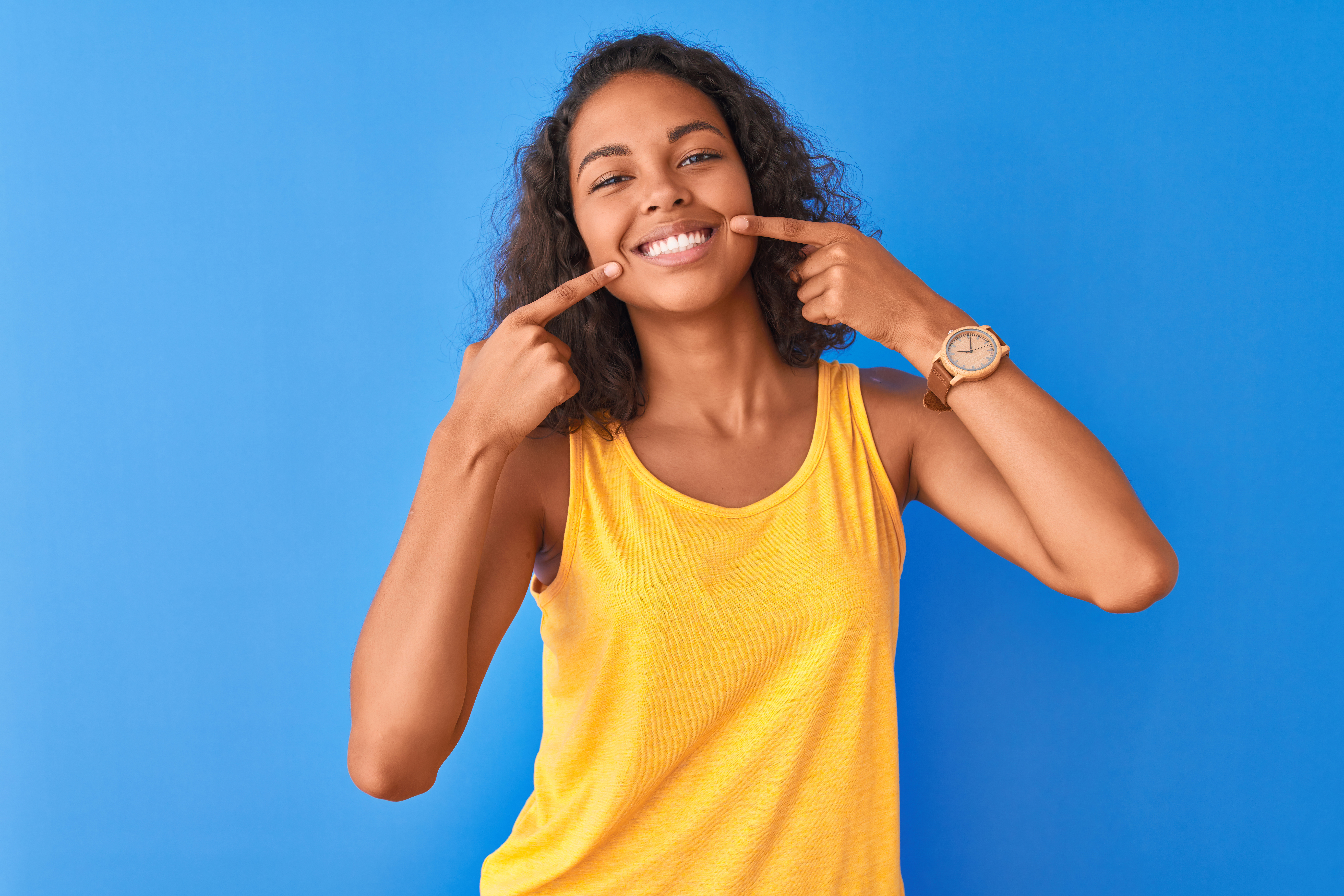 Young brazilian woman wearing yellow t-shirt standing over isolated blue background smiling cheerful showing and pointing with fingers teeth and mouth. Dental health concept.