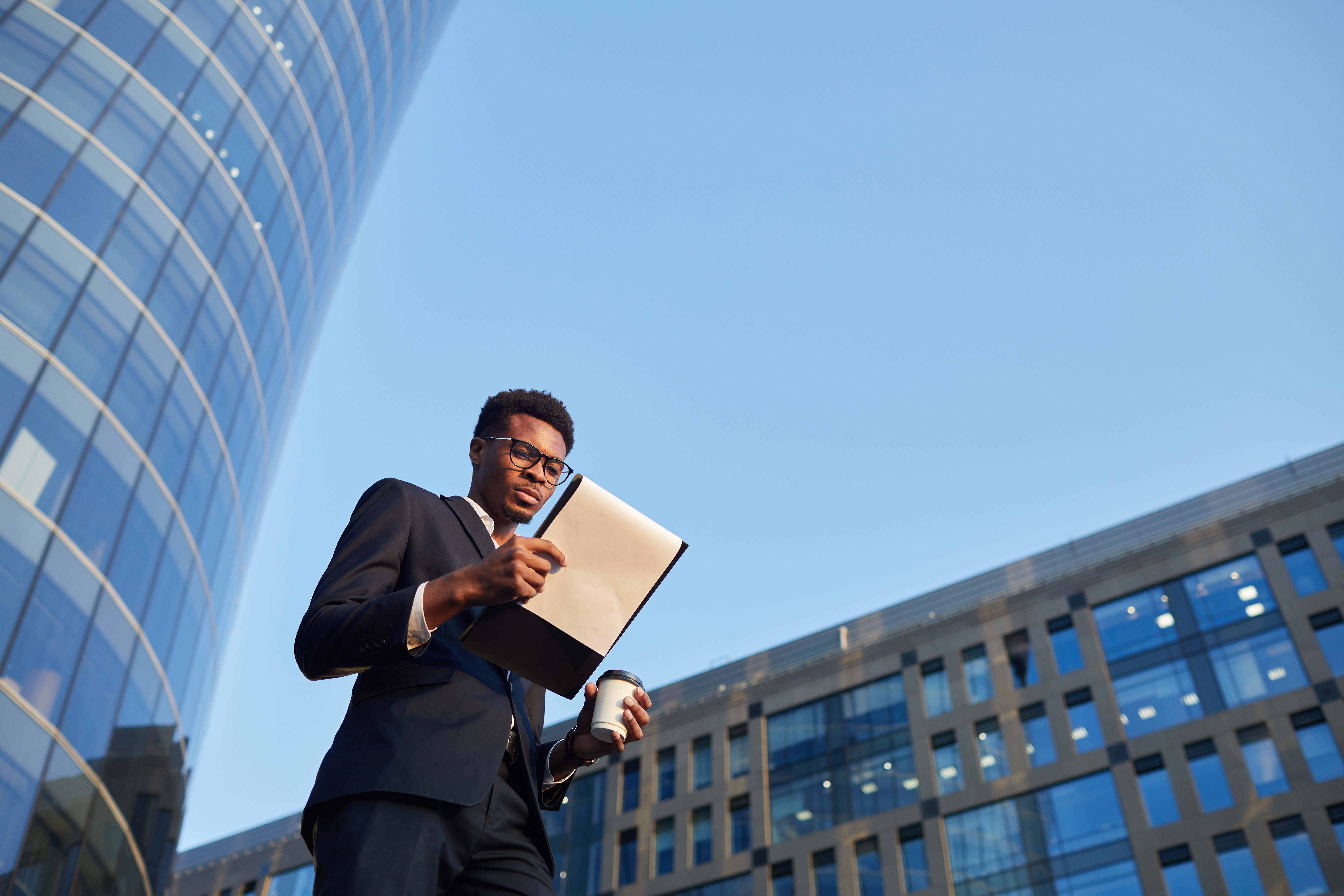 Below view of thoughtful young black businessman in elegant suit walking over business district and examining papers on move