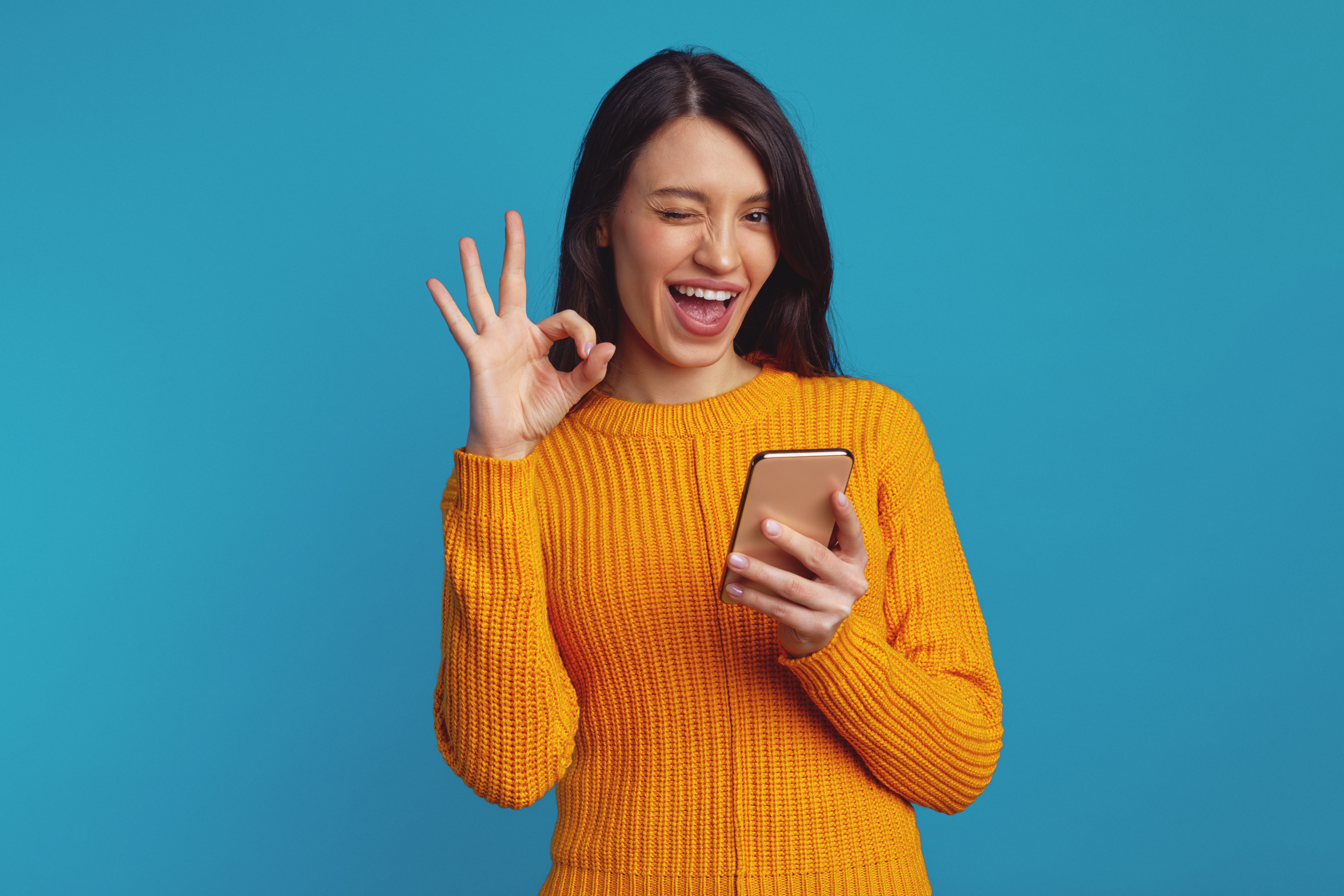 Young beautiful woman in orange sweater showing ok gesture while using smartphone